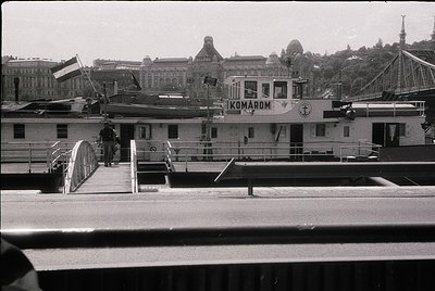 Black-and-white ferry docked at Budapest’s Danube River with "Ikomarom" branding, mid-20th century. Historic cityscape of dom...