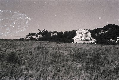 Vintage black-and-white aerial view of a lone wind turbine in a grassy field, surrounded by rocky terrain and sparse forest. ...