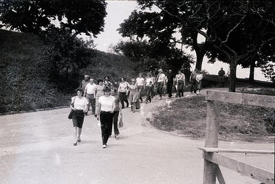 Group of people walking along a tree-lined path, likely a public park or promenade. Mid-20th century clothing (1950s–1960s) s...