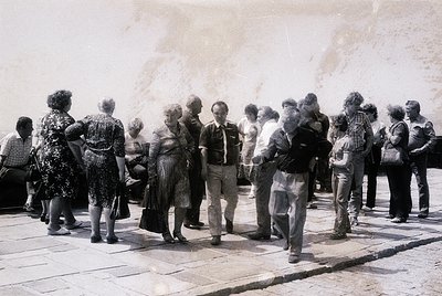 Group of 18+ people in 1960s-70s streetwear—women in patterned dresses, men in suits/vests—standing on paved plaza near concr...