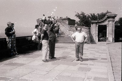 Black-and-white snapshot of a mid-20th-century gathering at a grand stone staircase with classical arches. A man in a white s...