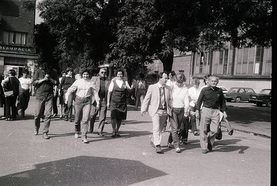 1970s street scene in a European city, likely Spain. Group of casually dressed pedestrians—men in suits, women in blouses and...