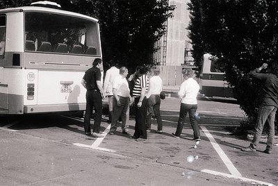 Group of five men in 1970s-era streetwear (striped shirts, bell-bottoms) standing near a white bus marked "100" in a parking ...