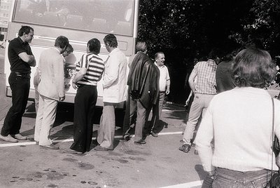 Black-and-white street scene: group of 7 adults in 1970s-era clothing (striped shirts, button-downs) standing in line at a bu...