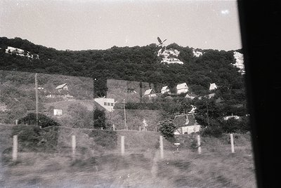 Mid-20th century rural landscape with scattered houses and wind turbines on rolling terrain. Foreground shows blurred roadway...