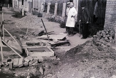 Mid-20th century street scene showing two men in formal attire observing a partially demolished brick wall and exposed founda...