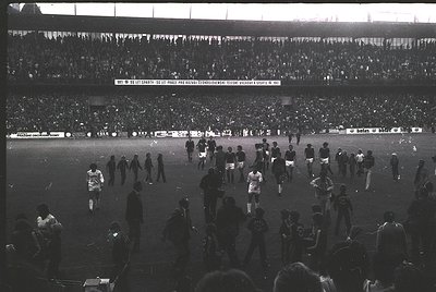 Black-and-white stadium scene capturing post-match chaos, 1960s-70s. Crowd surges onto field; players and spectators mix amid...