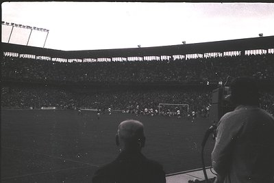Vintage black-and-white shot of a packed stadium during a soccer match, likely from the 1950s–1960s. Crowds fill tiered seati...