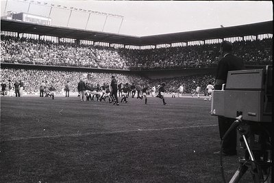 Vintage black-and-white shot of a packed stadium during a mid-20th-century football match. Players in shorts and boots engage...