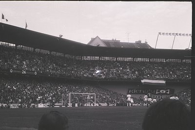 Vintage black-and-white stadium shot showing packed stands with dense crowd. Players in white uniforms near goalpost; visible...