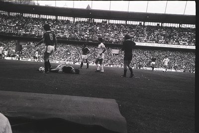 Black-and-white shot of a 1970s soccer match in a packed stadium, featuring a fallen player and teammates reacting. Uniforms ...