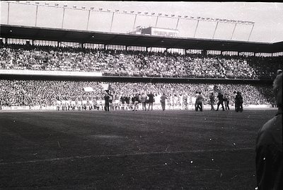 Vintage black-and-white stadium shot showing packed tiered seating with dense crowds. Players in vintage uniforms line up on ...