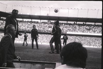 Black-and-white shot of a pre-game soccer ritual at a large stadium, likely 1960s–1970s. Players in striped jerseys toss a ba...