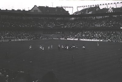 Black-and-white stadium shot showing a packed 1950s-era football match with tiered seating and flag-lined rooftops. Players a...