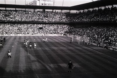 Classic black-and-white shot of a packed stadium during a mid-20th century soccer match. Players in vintage striped jerseys e...
