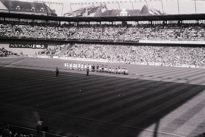 Vintage stadium shot featuring a large crowd in tiered seating, likely from the 1950s–1960s. Central field shows a marching b...