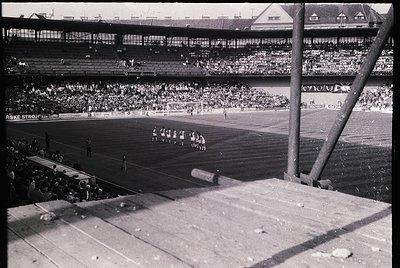 Mid-20th century stadium scene with packed stands, likely European. Players in white uniforms on a grass field, surrounded by...