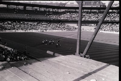 Mid-20th century stadium scene with multi-tiered concrete seating filled with spectators. A cycling race unfolds on the track...