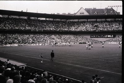Vintage black-and-white stadium shot showing mid-20th century football match. Multi-tiered seating packed with spectators, wi...