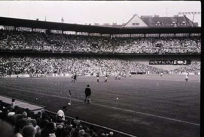 Vintage black-and-white stadium shot showing a packed 1960s-era football match. Multi-tiered seating filled with spectators, ...