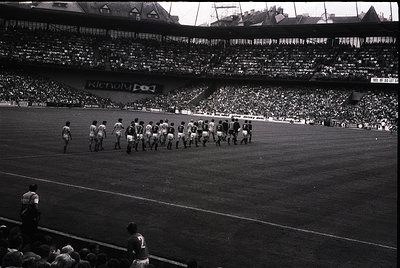 Black-and-white stadium shot of a pre-match football (soccer) team walkout, likely 1960s–1970s. Players in white kits line up...