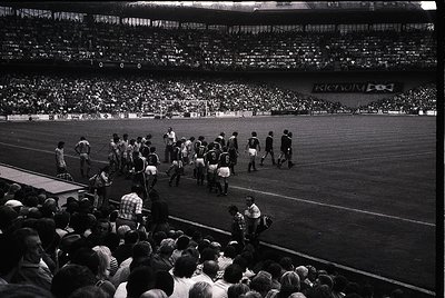 Black-and-white shot of a packed stadium during a pre-match moment, likely 1960s–1970s. Players in vintage striped jerseys an...