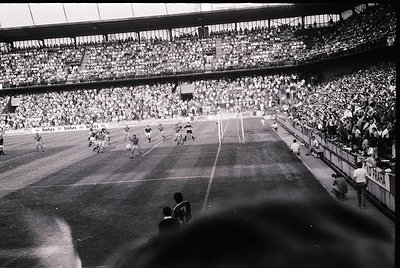 Vintage black-and-white stadium shot capturing a packed 1960s European football match. Players in white and dark jerseys cont...