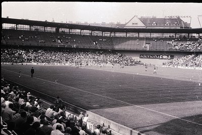 Vintage black-and-white stadium shot showing a packed 1960s-era football match. Multi-tiered stands filled with spectators, p...