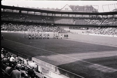 Black-and-white shot of a mid-20th-century stadium with players in formation, likely pre-game warm-up. Multi-tiered seating w...