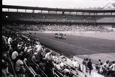 Vintage black-and-white photo of a packed stadium during a soccer match, featuring tiered seating and a large crowd. Players ...