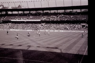 Vintage black-and-white stadium shot showing a packed 1960s-era football match. Players in striped jerseys engage in action o...