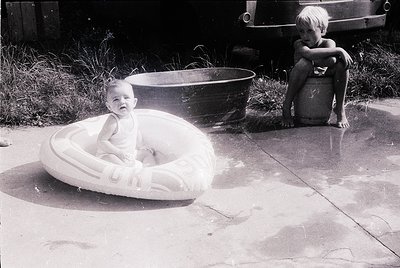 Mid-20th century backyard scene: infant in a white inner-tube on concrete, older child seated on a barrel. Classic 1950s–60s ...