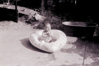 Vintage black-and-white photo of an infant seated in a large, inflatable pool ring on a concrete patio. Surrounding elements ...