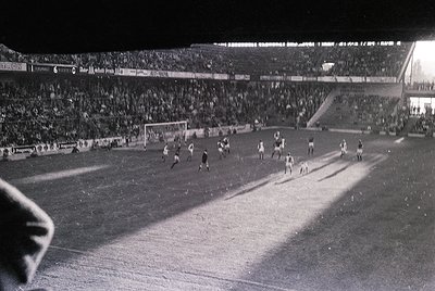 Black-and-white shot of a packed stadium during a 1960s–1970s football match, likely in Europe. Crowds fill tiered stands und...