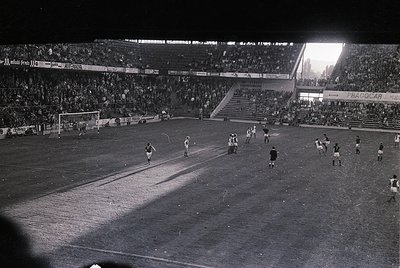 Black-and-white nighttime soccer match in a large stadium, featuring a packed crowd under floodlights. Players in dark unifor...
