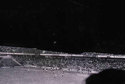 Black-and-white night shot of a packed stadium, likely a European football match from the 1960s–1970s. Crowds fill tiered sta...