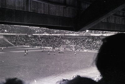 Black-and-white shot of a packed stadium during a mid-20th-century football match. Crowds fill tiered stands, with players an...
