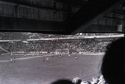 Black-and-white stadium shot capturing a mid-20th-century football match. Crowded stands with sparse advertising (e.g., "Tech...