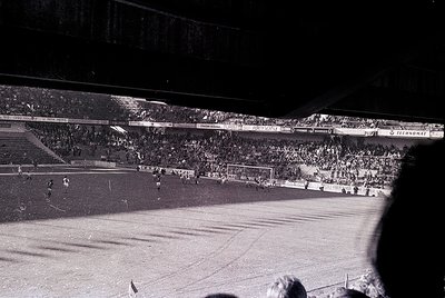 Vintage black-and-white shot of a packed stadium during a soccer match, likely 1960s–1970s. Crowds fill tiered stands under f...