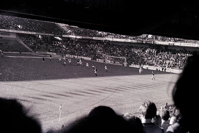 Black-and-white shot of a packed stadium during a soccer match, likely 1960s–1970s. Crowd fills tiered stands, with visible a...