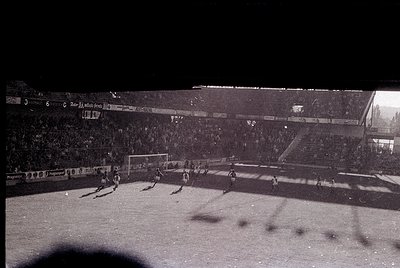 Vintage black-and-white shot of a packed stadium during a nighttime football match. Crowds fill tiered stands under floodligh...