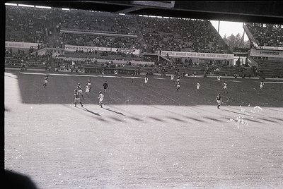 Black-and-white shot of a packed stadium during a mid-20th century football match. Visible banners advertise brands like "Voo...