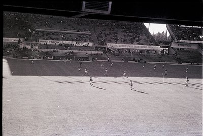 Vintage black-and-white shot of a packed stadium during a soccer match, featuring midfield action with players in motion. Adv...