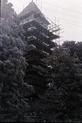 Multi-tiered pagoda under restoration, surrounded by dense foliage. Scaffolding covers each level, indicating preservation wo...