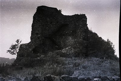 Ruined medieval stone fortress perched on rocky hillside, surrounded by sparse vegetation. High-angle vintage photograph capt...