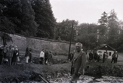 Mid-20th century black-and-white photo of a rural gathering near a stone wall and small wooden structure. Central figure in s...