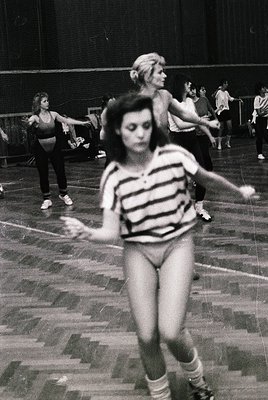 Vintage black-and-white gym class scene, 1960s-70s. Women in form-fitting leotards and knee-high socks practice dance or gymn...