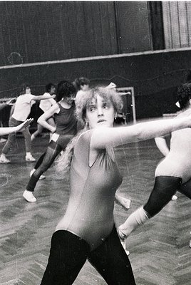 Black-and-white shot of a dance studio session, likely 1960s–1980s. Foreground dancer in sleeveless top and tights, mid-movem...