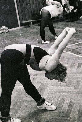 Black-and-white gymnastics warm-up in a 1960s–70s indoor facility. Female athlete in leotard and sneakers performs a forward ...