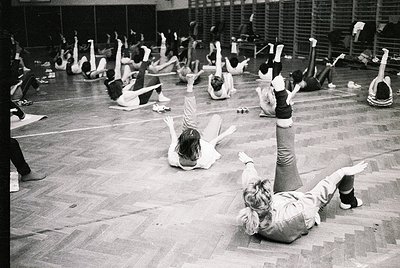 Group aerobics class in an indoor gym, likely 1970s–1980s. Participants perform floor exercises in unison, wearing leotards a...